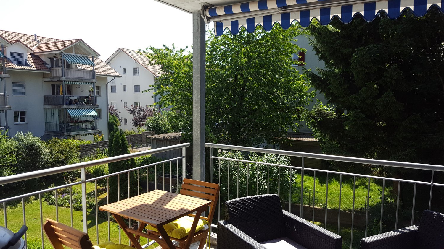 Balcony with tables and chairs, awning, fenced with metal railings, overlooking grass field