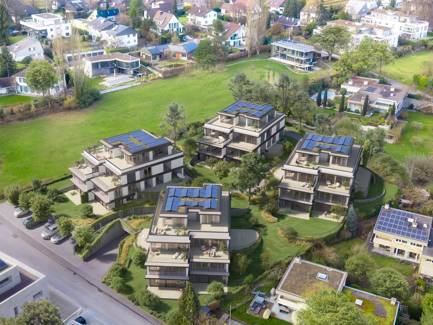 Aerial view of a residential complex with multiple modern, multi-story buildings surrounded by lush greenery and landscaping. The buildings feature balconies, terraces, and solar panels on the roofs. There are also parking spaces visible on the grounds.