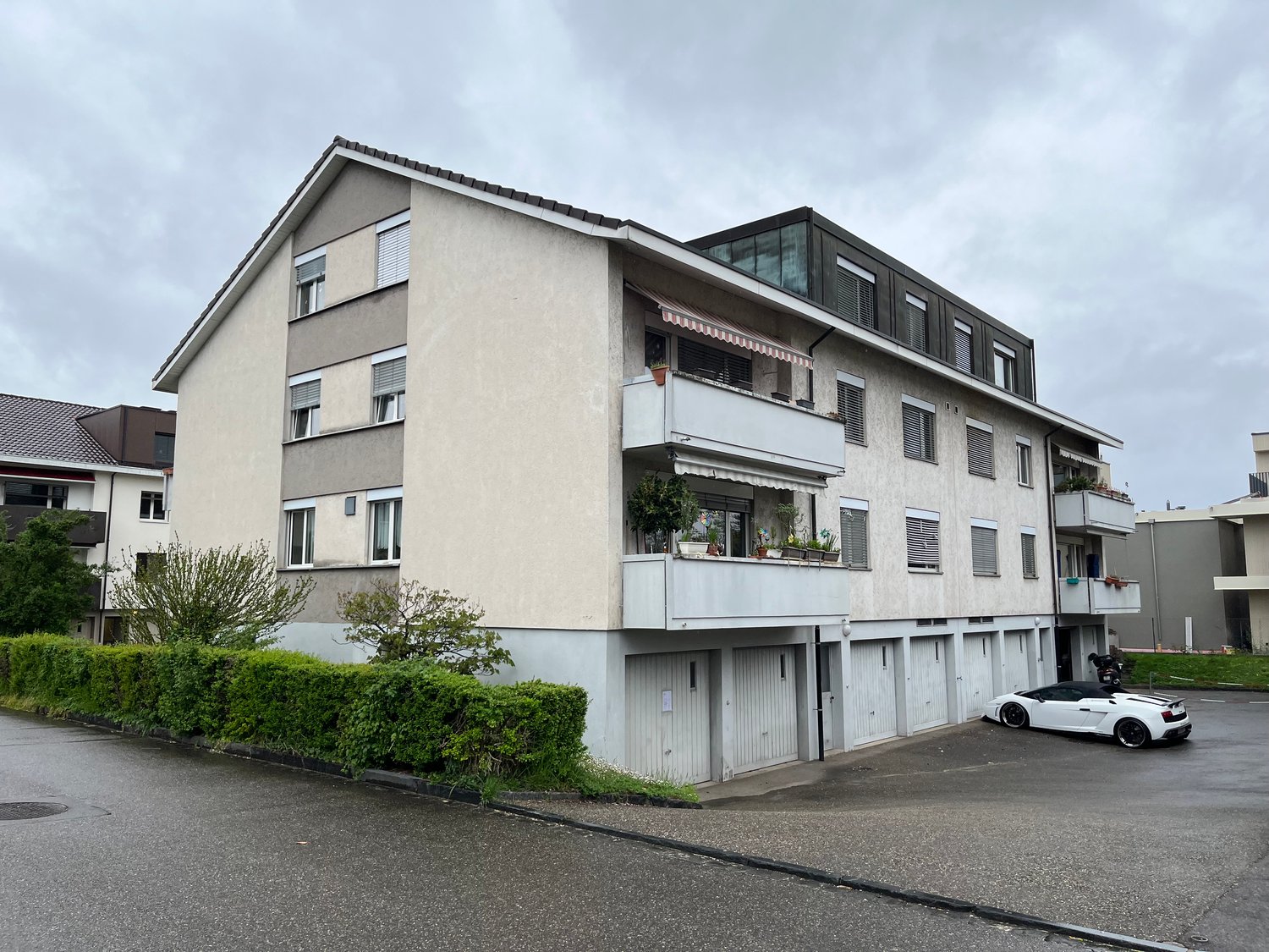 Multi-story apartment building, beige exterior, multiple balconies, potted plants, three garage doors, car parked in front