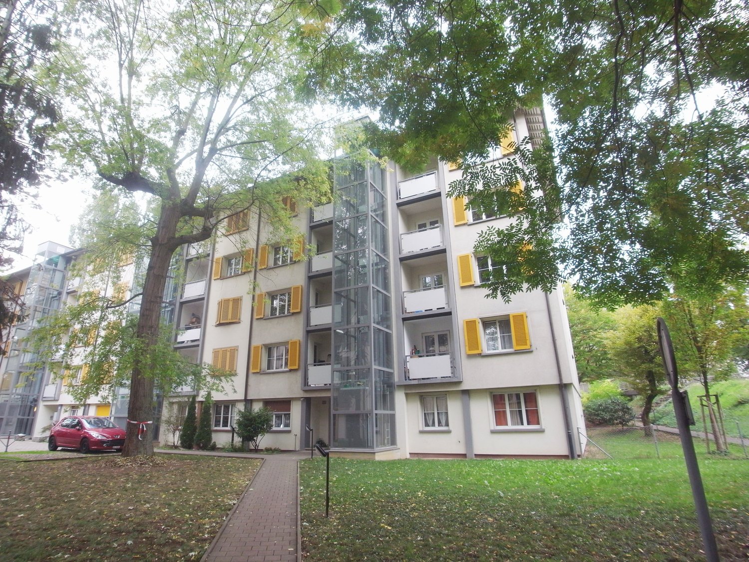 Residential building, white and yellow paint, yellow shutters, glass elevator, trees, walkway, car parked