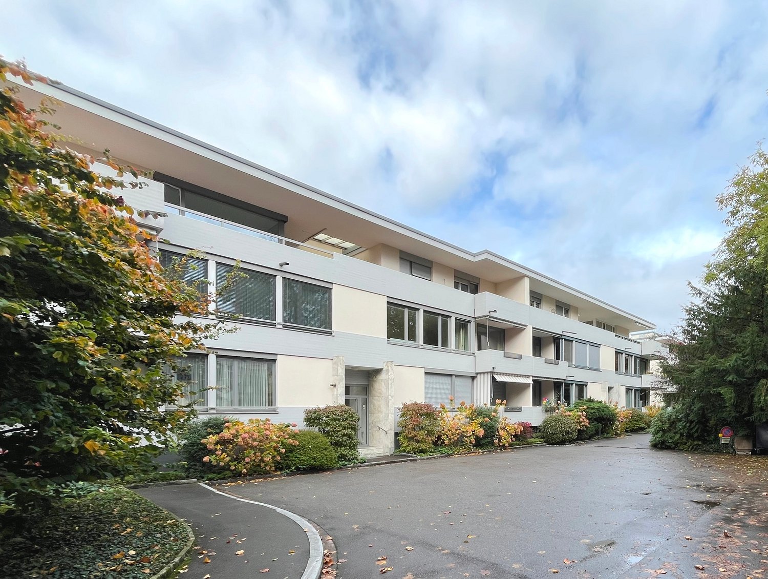 3 story building, white and gray exterior, large windows, balconies, entrance door, road, shrubs and trees