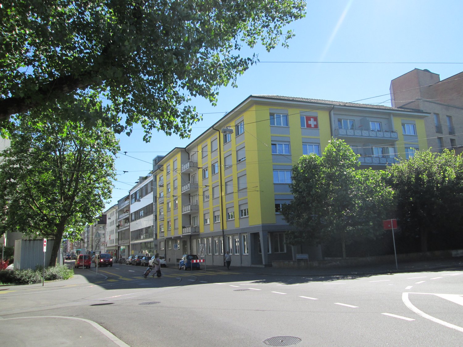 Yellow residential building with many windows, balconies and cross symbol, several other buildings on the street, streetlight, people walking, cars parked on the street.