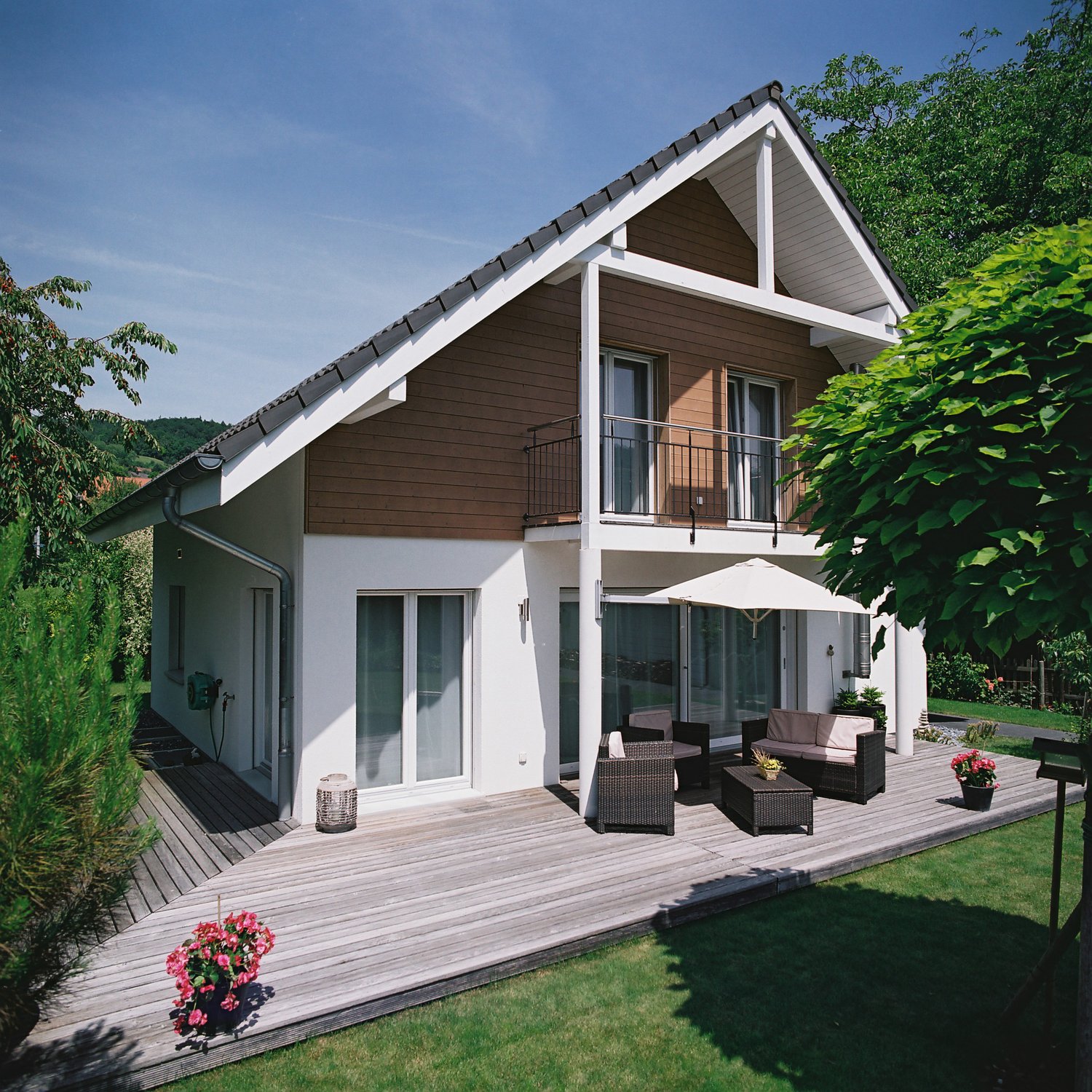 Detached house, 2 stories, brown and white, glass windows, wooden deck, chairs and table, umbrella, flower pots