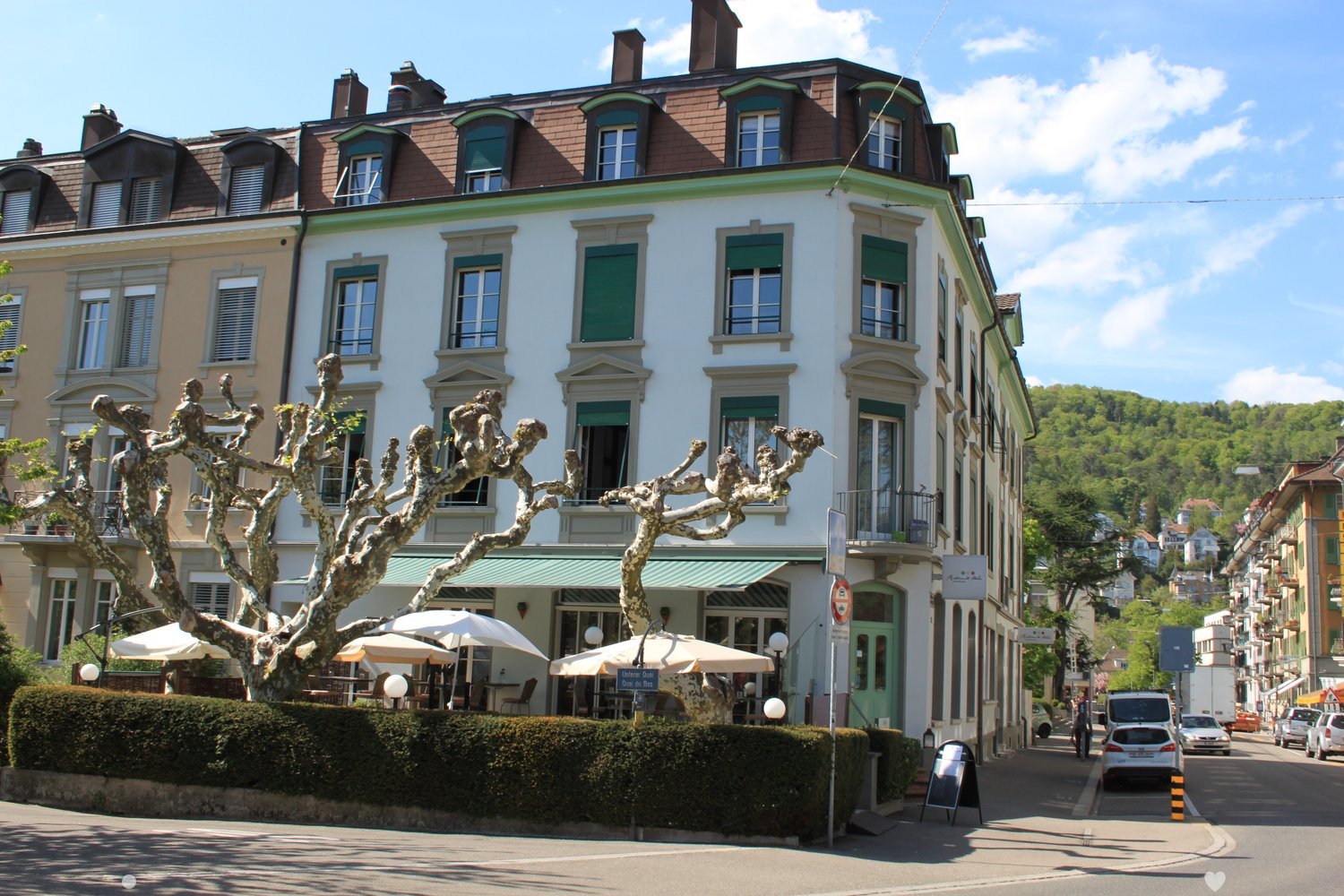 Large building with multiple stories, windows, and a green awning in front, outdoor tables and umbrellas in front, street scene with trees, parked cars, and people walking