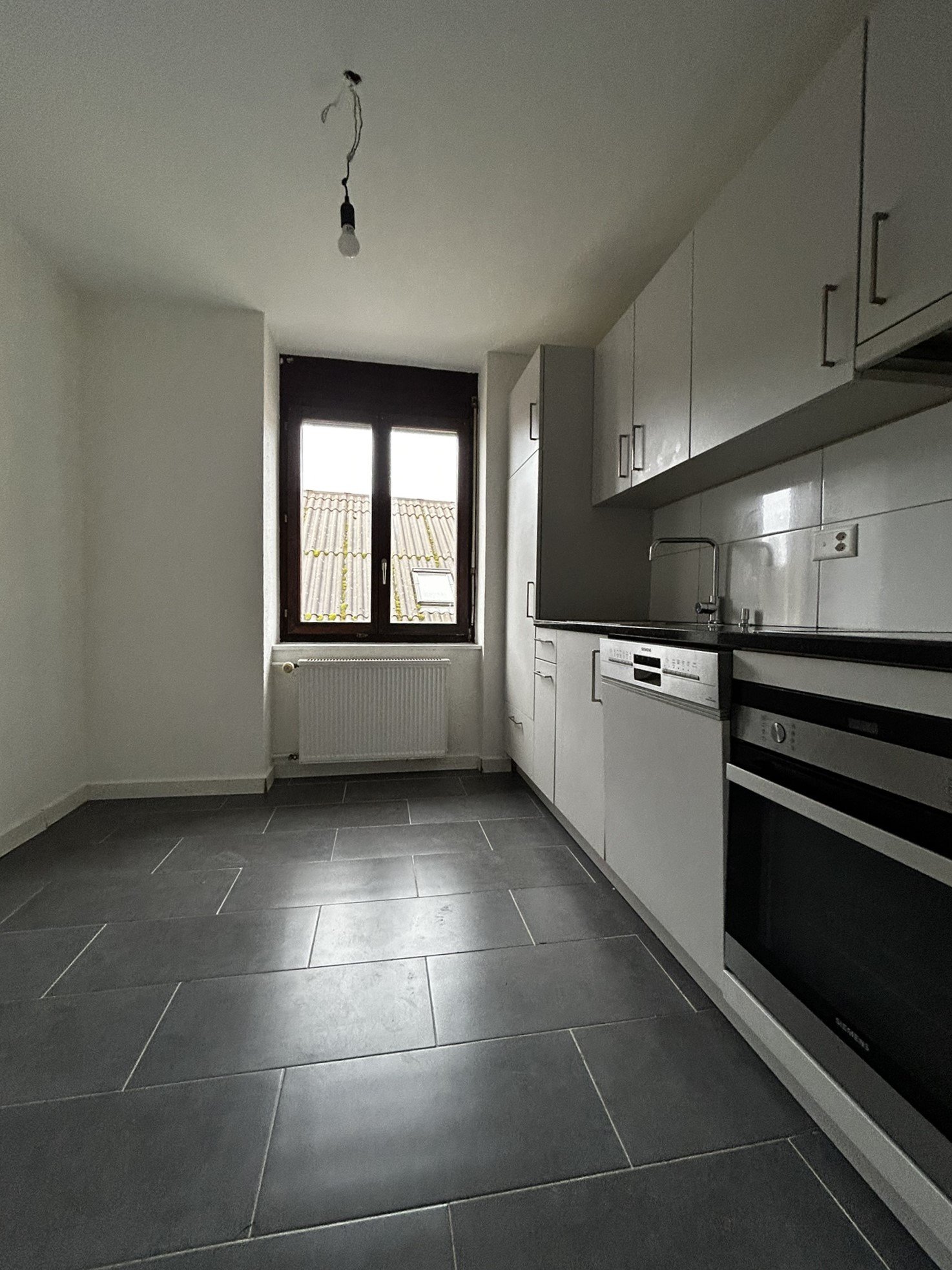 Spacious kitchen with modern white cabinets, black countertops, and a dishwasher. The room has a large window providing natural light. The floor is covered with grey tile.