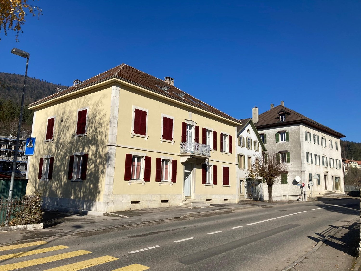 A multi-story yellow building with red shutters, located on a street with other buildings in the background. The building appears to be a residential or commercial structure in a town or city setting.