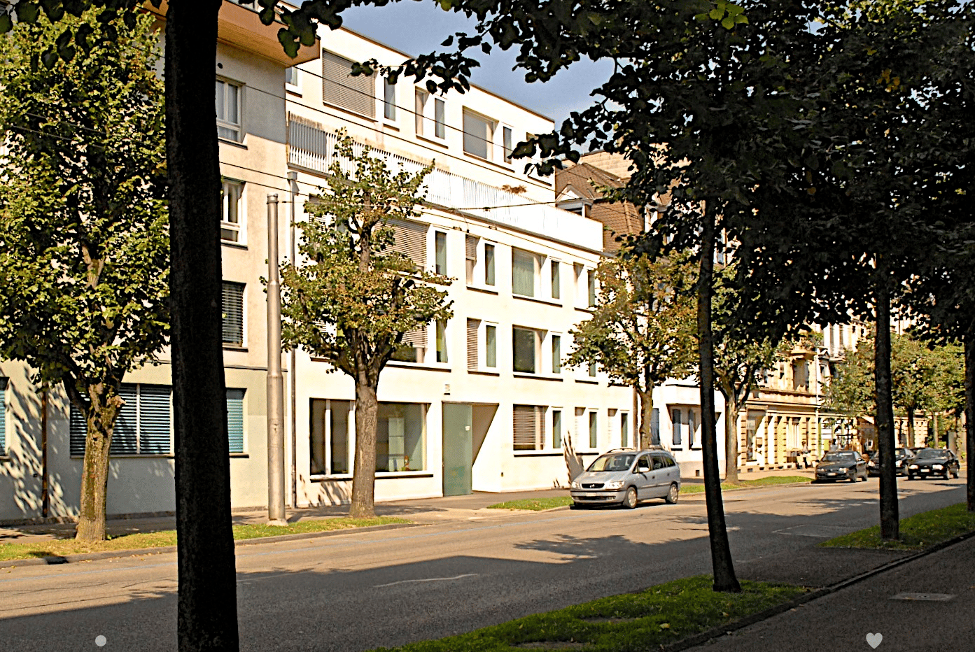 Building along a street with cars parked and cars passing by, multiple windows and green door, trees along the street.