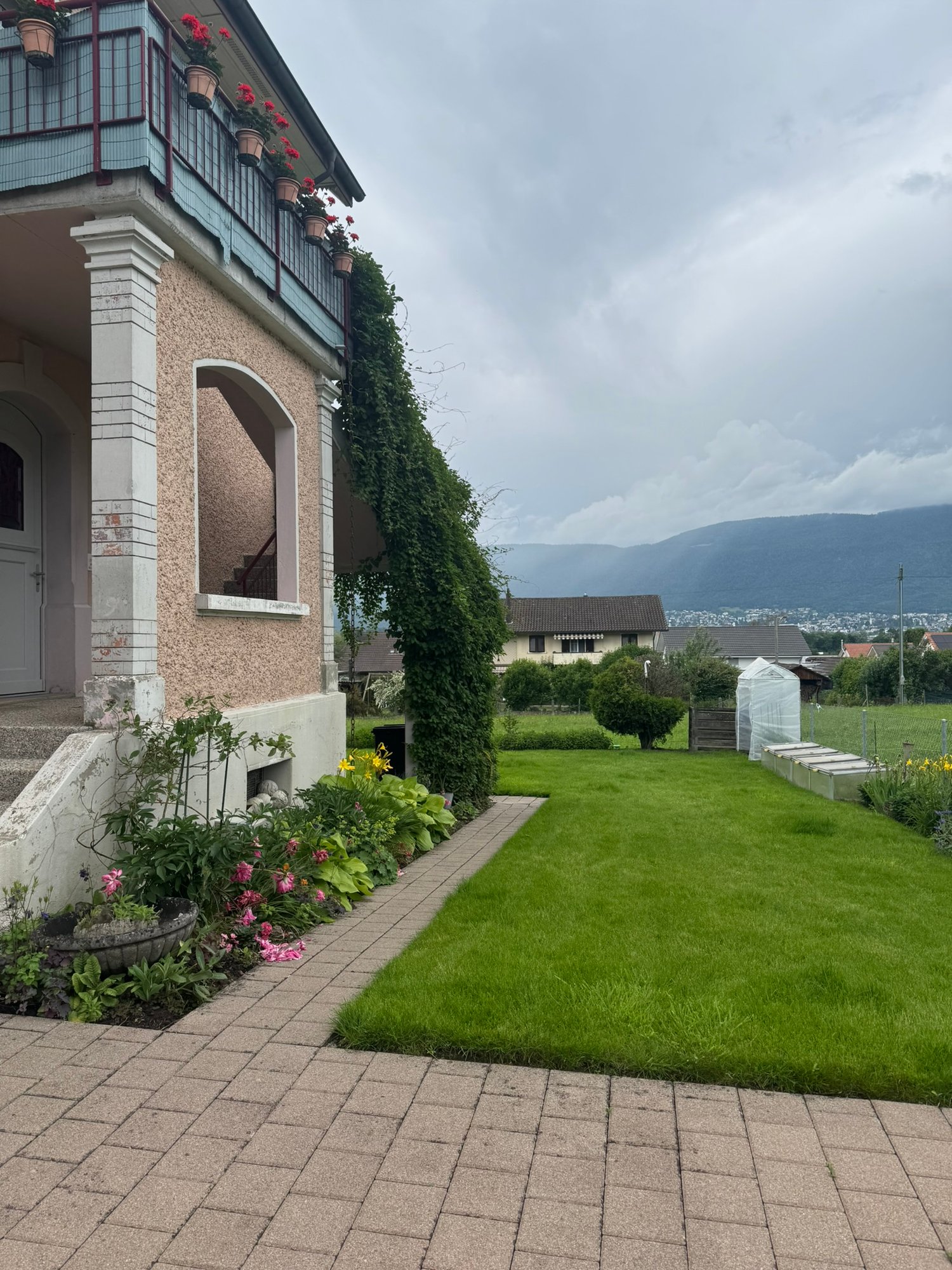 Two-story building with arched windows, balconies, and hanging flower baskets. The building is surrounded by a well-maintained garden with a grassy lawn, flower beds, and a greenhouse. The background shows a mountainous landscape.