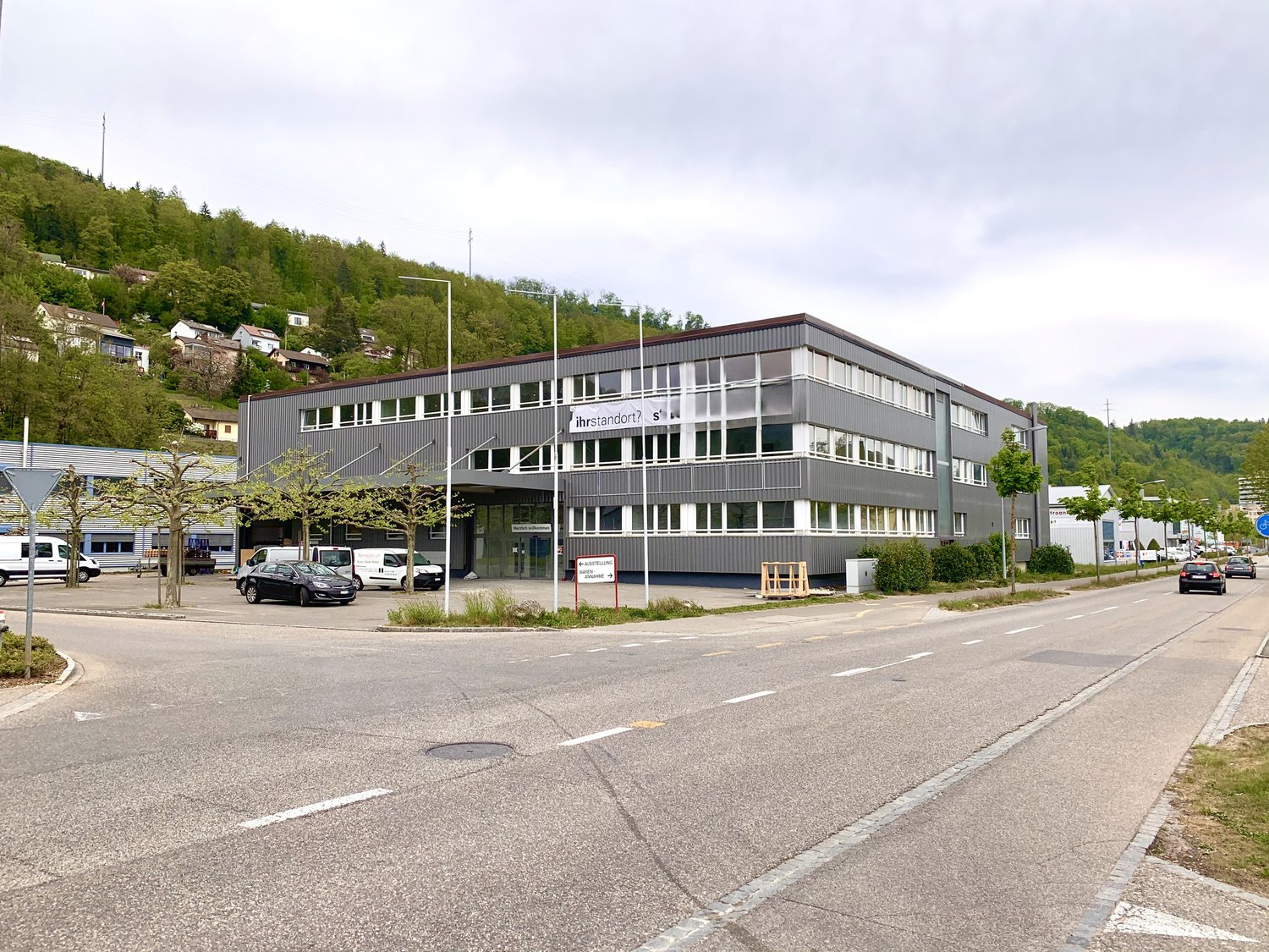 A multi-story commercial building with a gray exterior and large windows. The building appears to be located in a hilly, forested area with other smaller buildings and vehicles visible in the surrounding area.