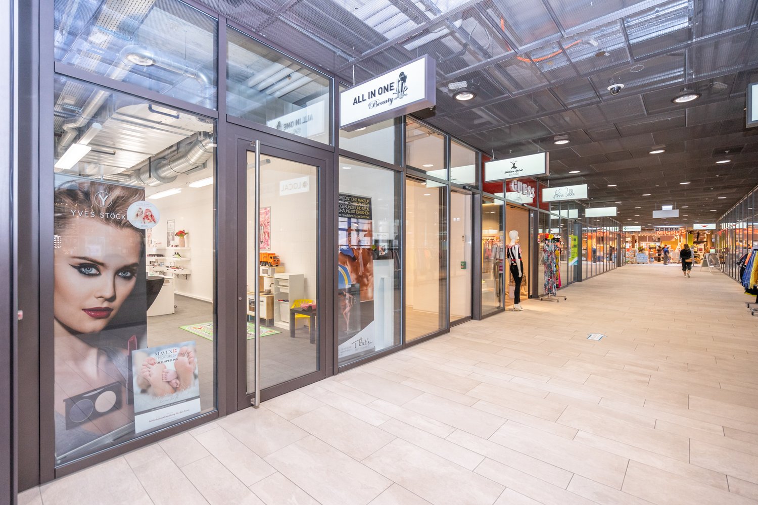 A modern shopping mall with glass walls, tiled floor, and several stores visible through the windows.