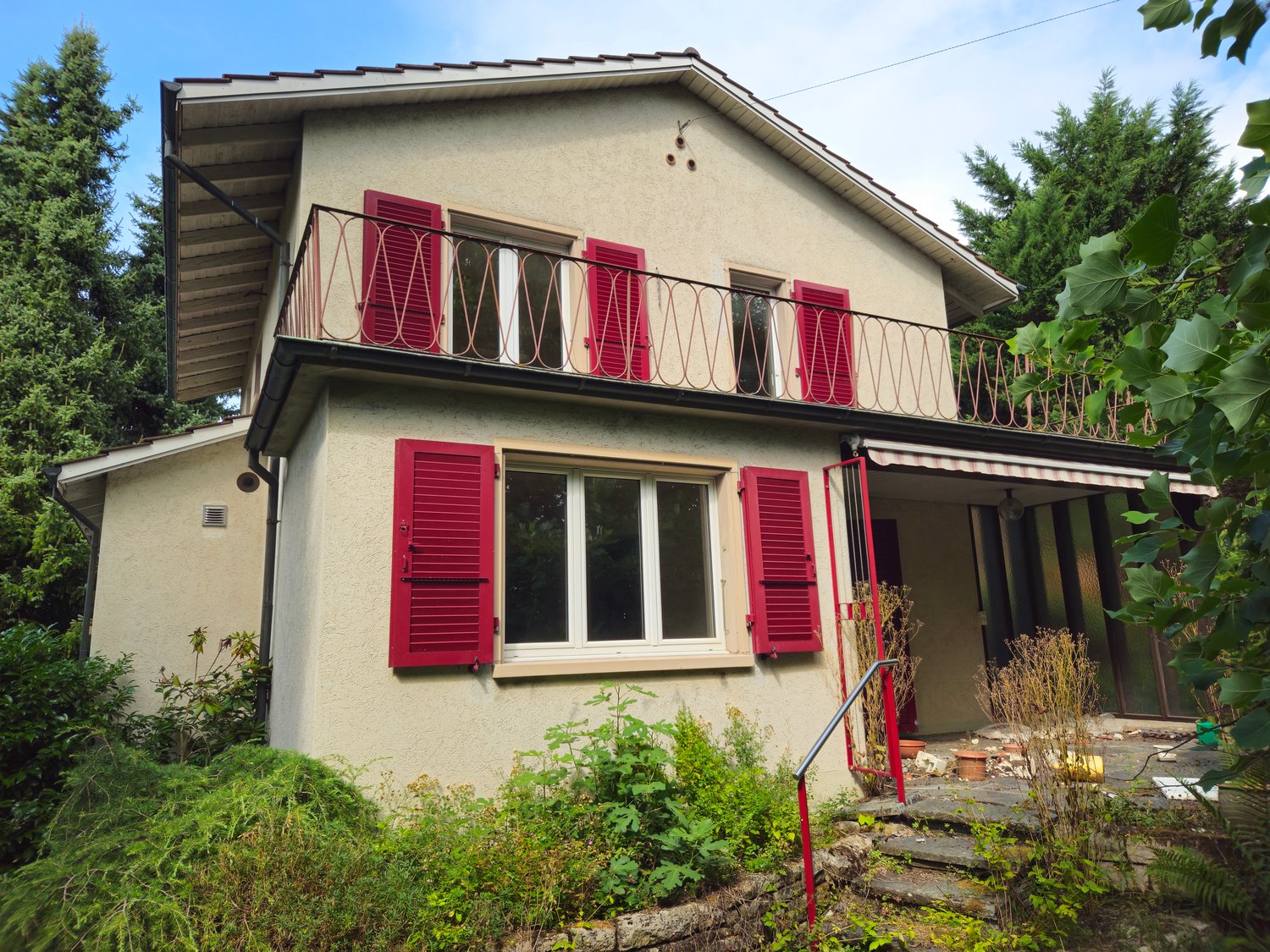 Two-story house with red shutters, beige walls, wooden terrace with a metal railing, awning over the entrance, and surrounded by greenery