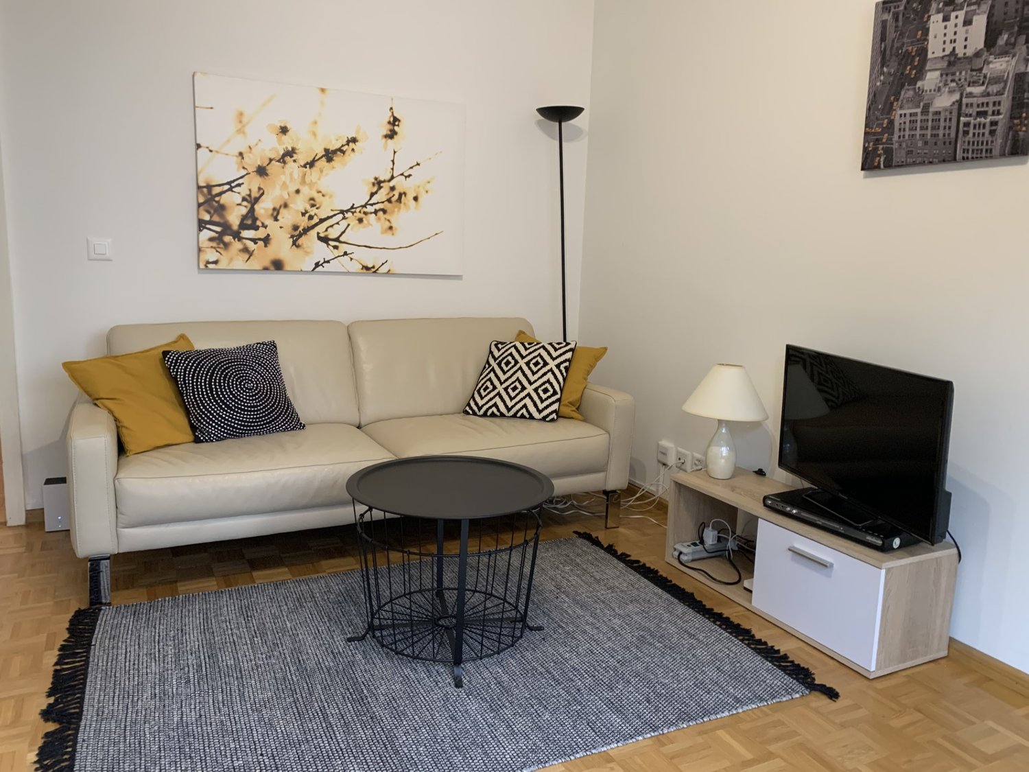 Beige sofa with patterned throw pillows, black and white coffee table, TV stand with TV, artwork on the wall, floor lamp, and a rug on the hardwood floor.