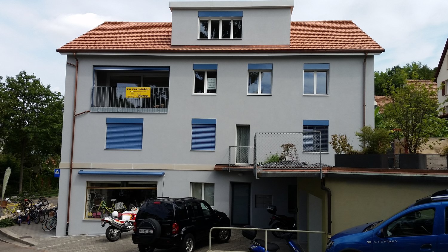 Modern two-story building with red roof, blue shutters, rental sign on the window, two cars parked in front.