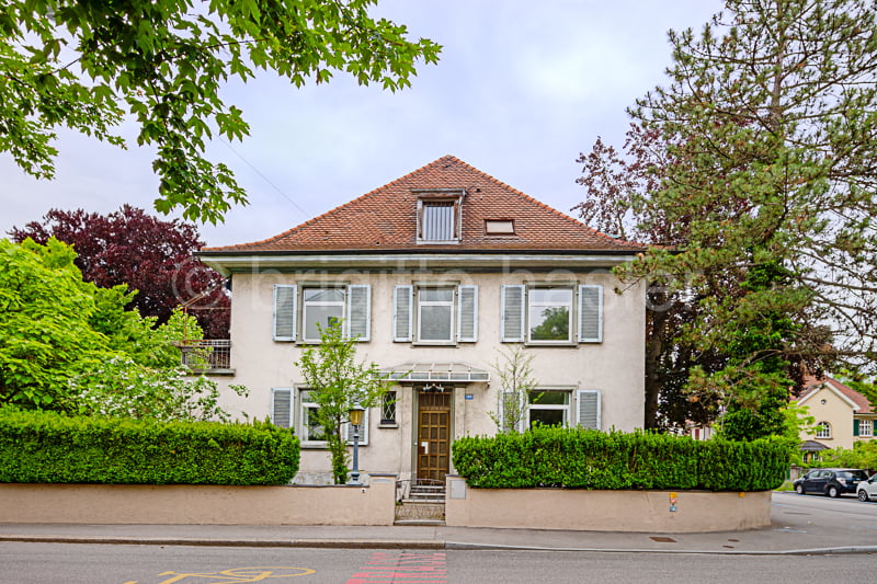Two-story residential building with a red tiled roof, white walls, and shuttered windows. The house is surrounded by greenery, including trees and hedges. There appears to be a balcony or terrace on the upper floor.