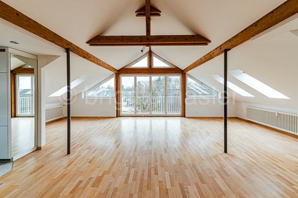 Spacious attic room with high ceilings, exposed wooden beams, and large windows providing ample natural light. The room has a hardwood floor and an open layout, suitable for a living or recreation area.