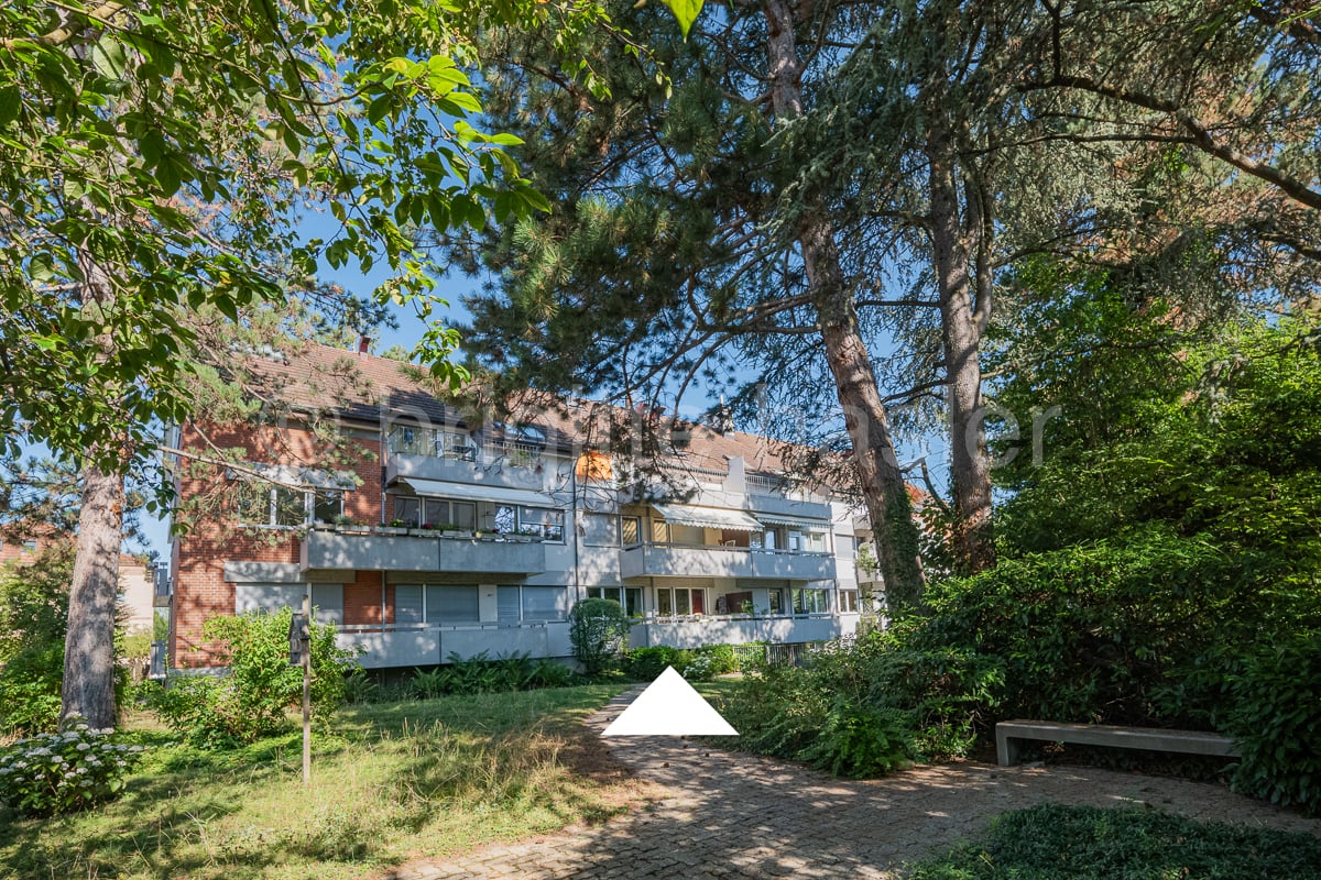 Multistory apartment building, brick exterior, large trees surrounding, grass garden, concrete pathway