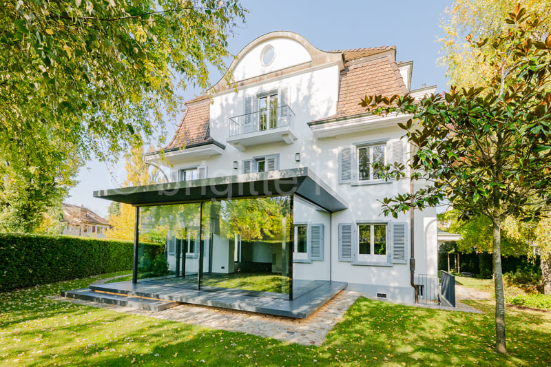 2 story house, white exterior, brown roof, terrace, glass walls, trees and bushes in the yard