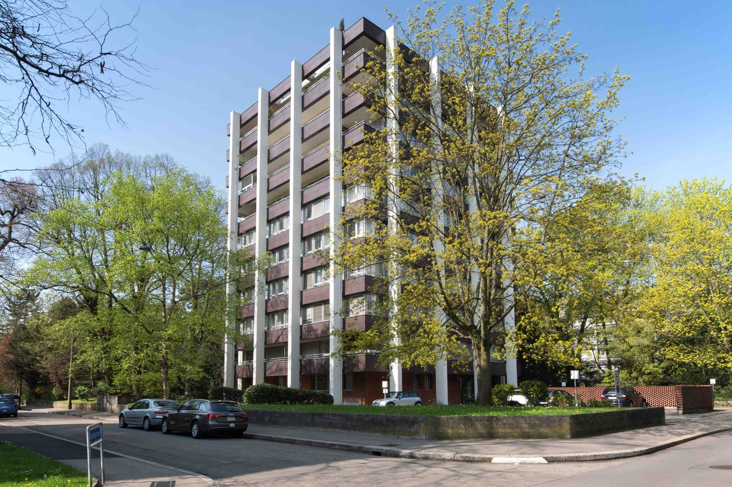 Modern apartment building with 5 floors, white and brown facade, large windows, several balconies, parking area, street with cars and trees