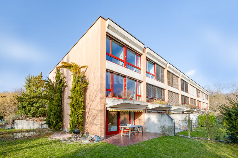 Modern building, pink exterior, red window frames, outdoor dining table, garden, small porch, shrubs and trees