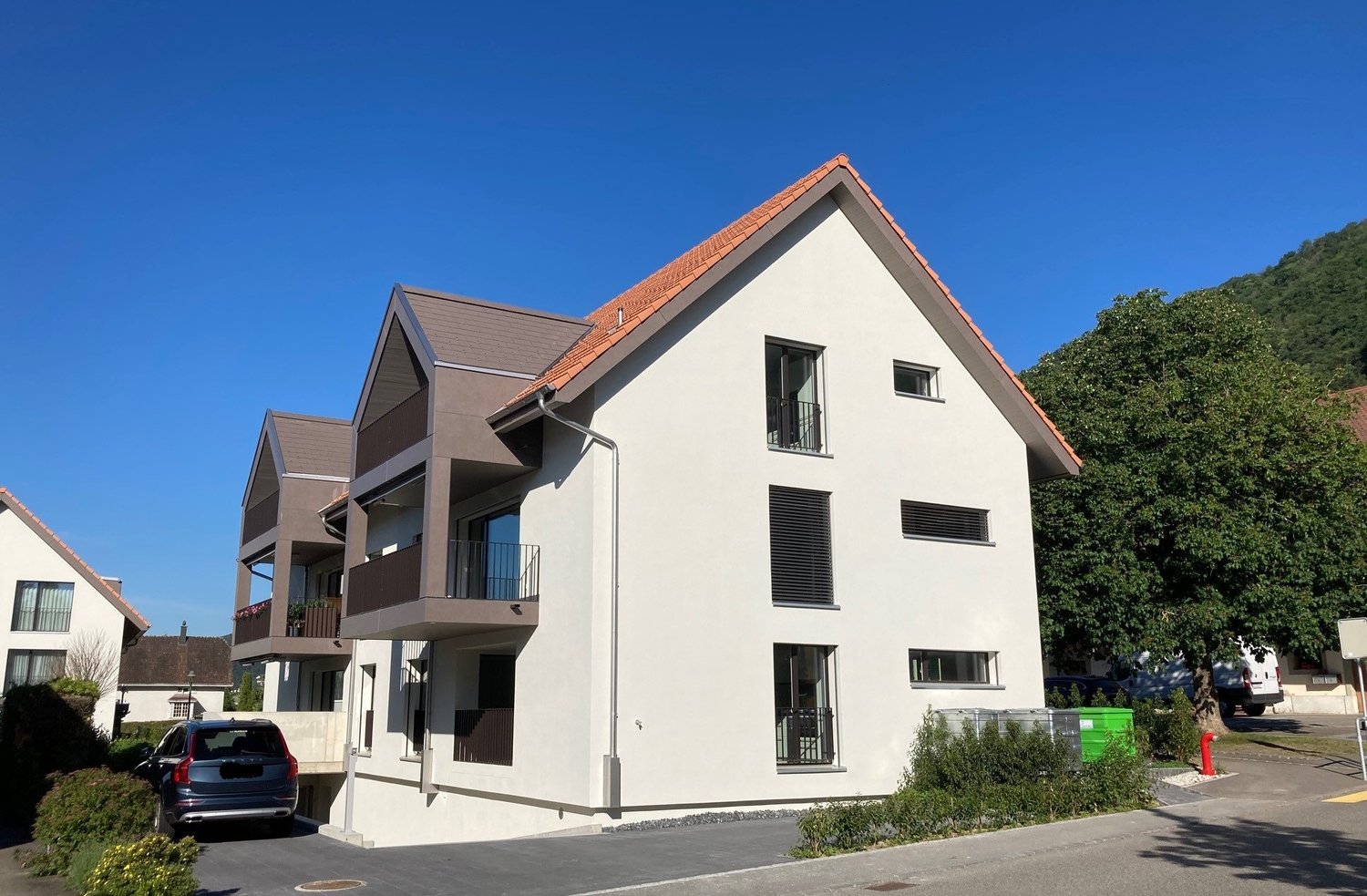 Three-story house, white walls, red roof, multiple windows, balconies, driveway, small garden