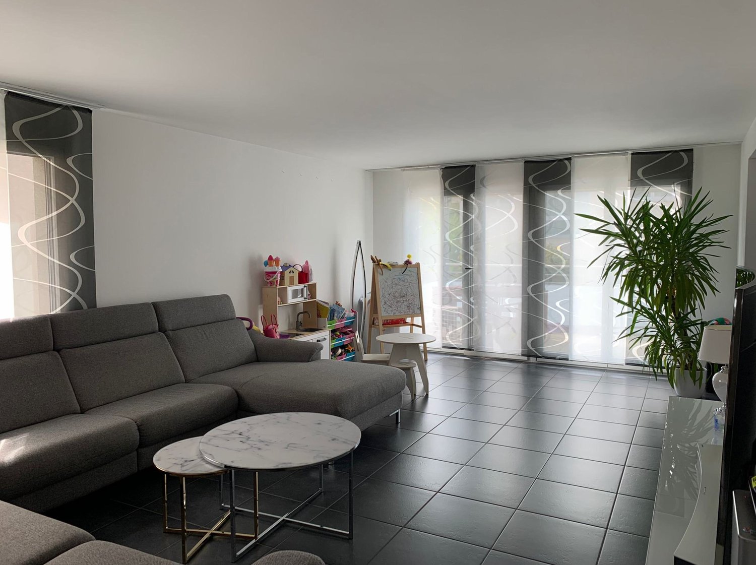 modern living room, white walls, grey couch, marble tables, tiled floor, plants, children's toys