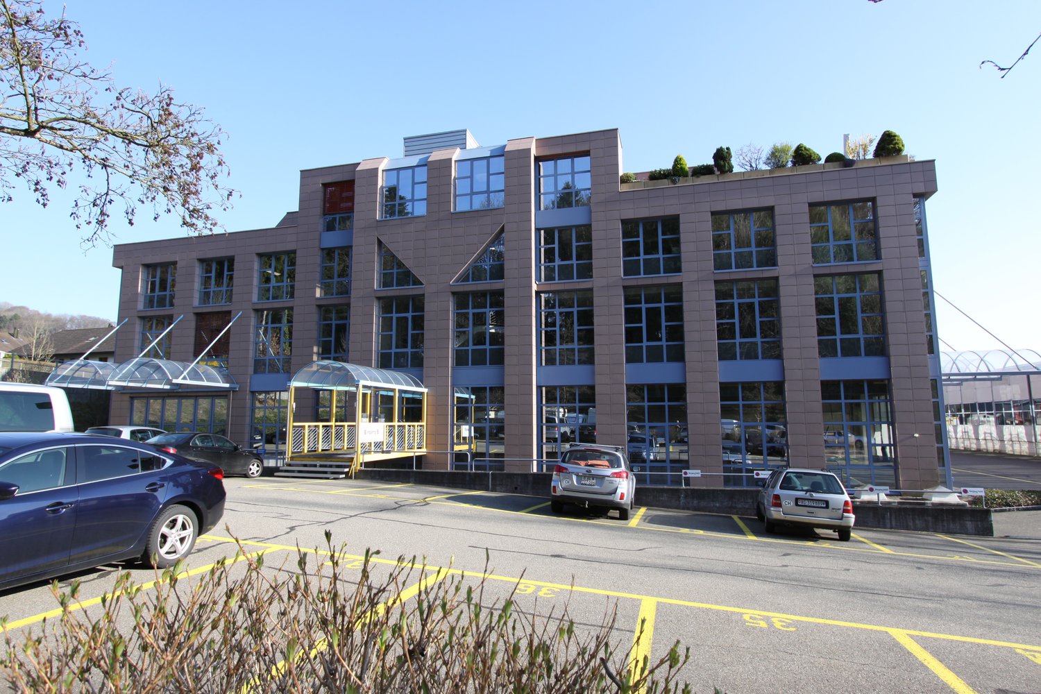 Large office building with many windows, roof terrace, large parking area in front of the building