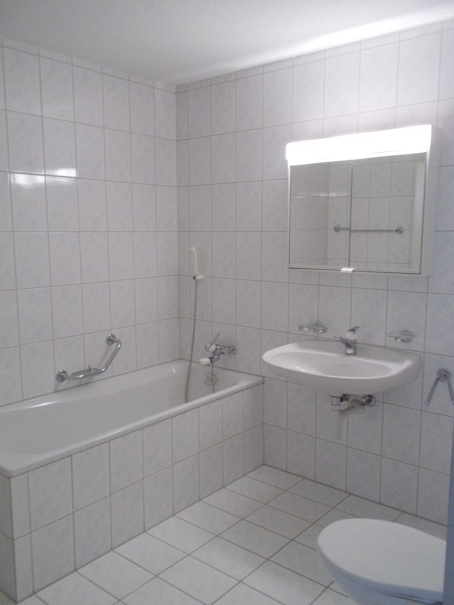 The image shows a bathroom with white tiled walls and floor. It has a bathtub, a sink with a mirror above it, and a toilet. The bathroom appears to be clean and well-lit.