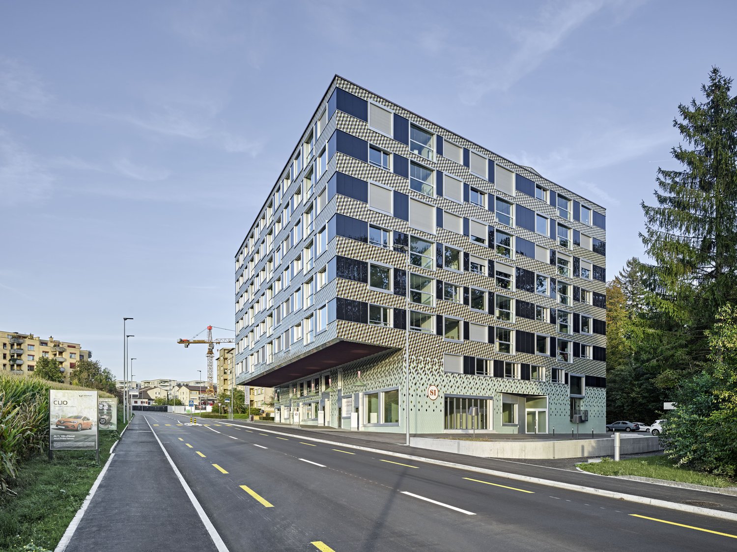 Modern apartment building with glass and metal facade, located on a road with trees and other buildings in the background.