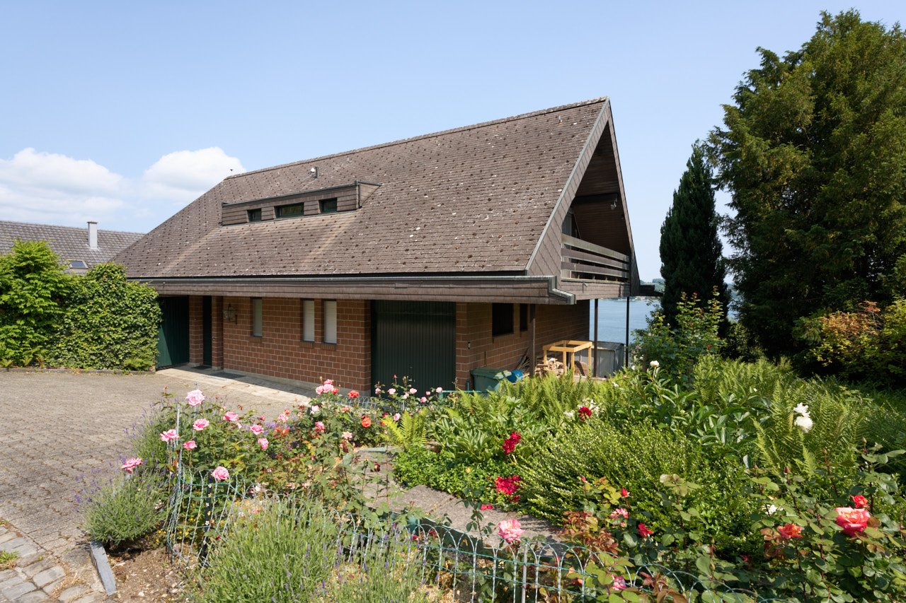 2-story brick house with a slanted roof, surrounded by a well-maintained garden with various flowers and plants. The house has a garage attached to the side.