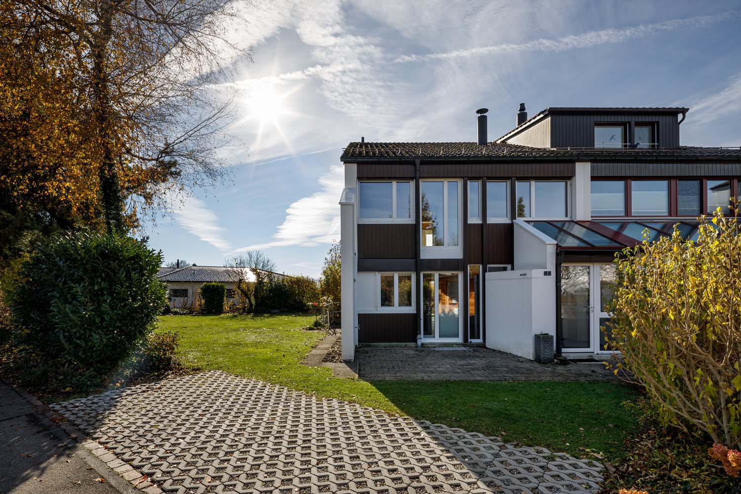 Modern two-story house with large windows, brown and white exterior, driveway with cobblestone pattern, lush garden, and clear blue sky with scattered clouds.