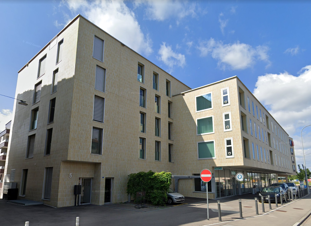 Modern multi-story building with beige tiles, multiple windows, and a flat roof. Located on a street with parked cars and a stop sign.