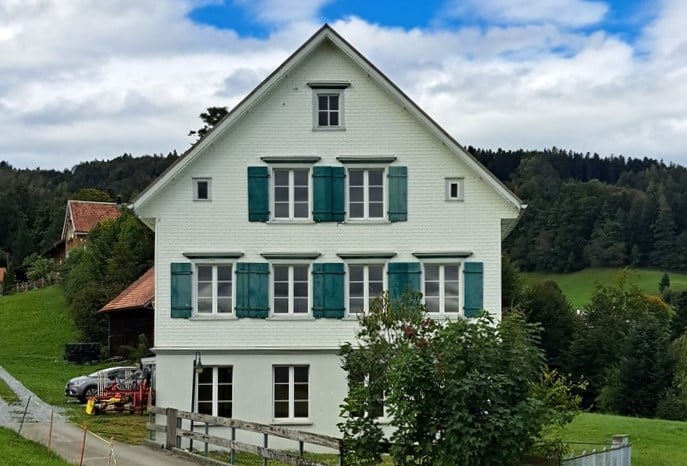 2-story white house with green shutters, surrounded by trees and a grassy field, located in a rural area with mountains in the background