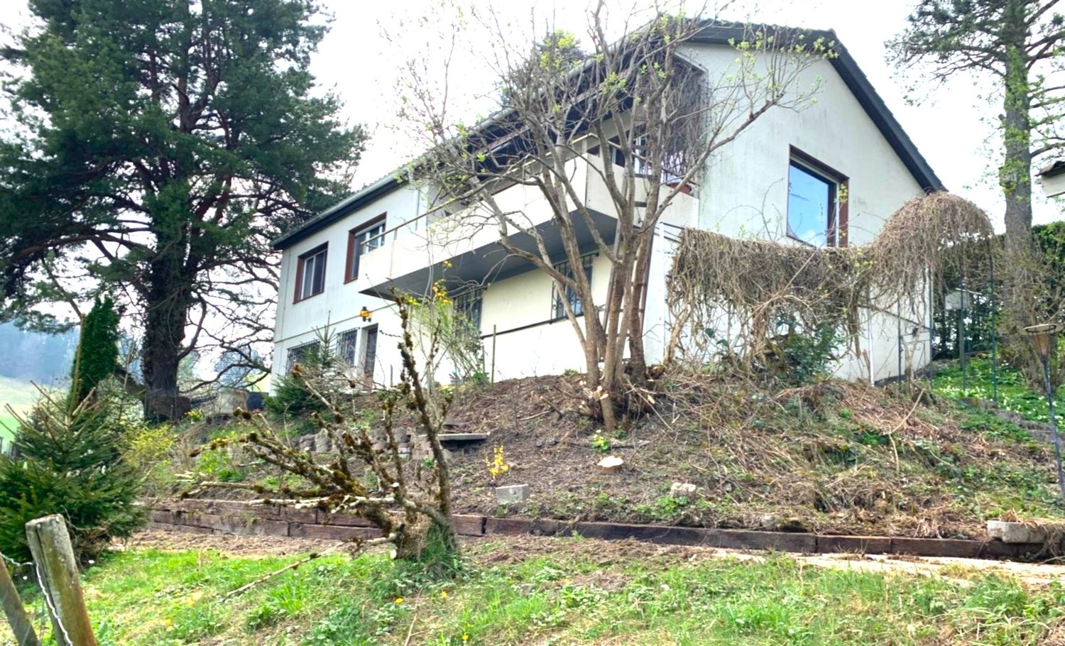 Two-story house with a balcony, surrounded by trees and vegetation. The house has a sloped roof and large windows. There is a raised garden area in front of the house.