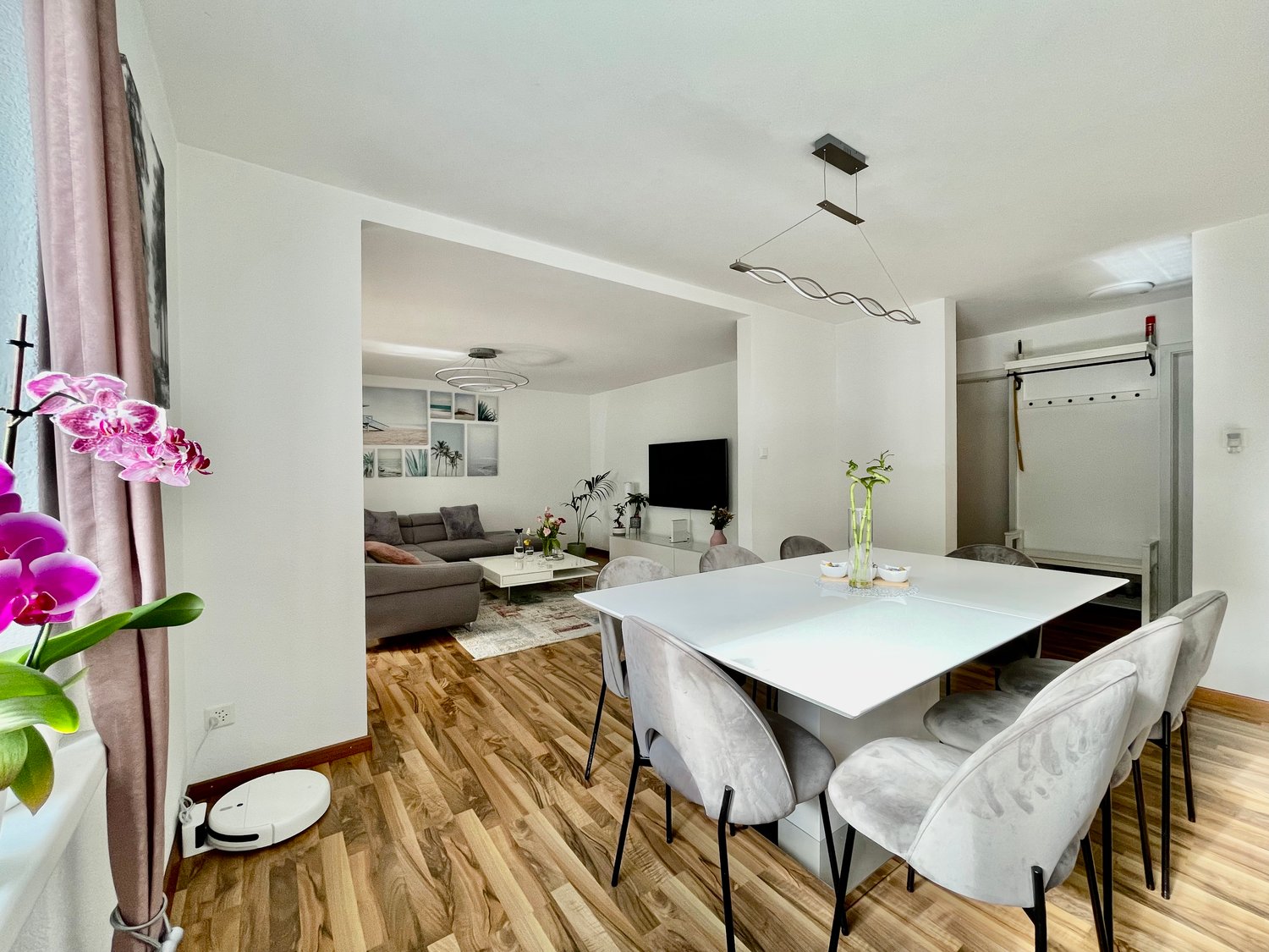 The image shows an open-concept living room and dining room area. The living room has a gray sofa and a TV mounted on the wall. The dining room features a white rectangular table with gray chairs. The floors are made of hardwood. The room is well-lit with