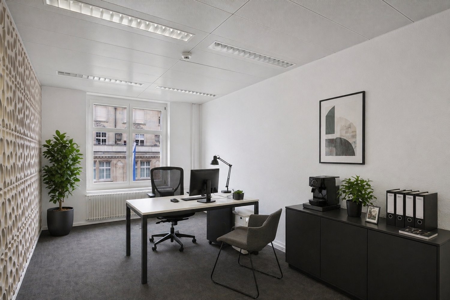 A modern office space with a desk, computer, and office chair. The room features a white wall, a radiator, and a window with a view of a building. There is a plant on the desk and a picture frame on the shelf. The room is empty and well-lit.