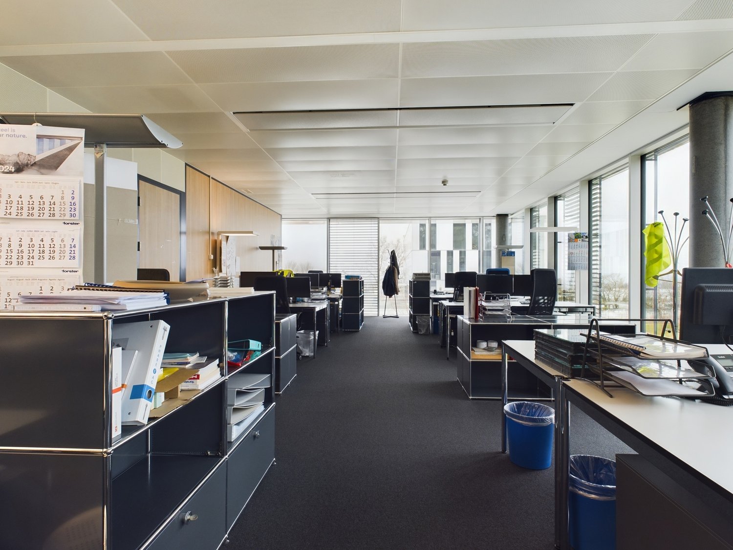 Open office space with cubicles, desks, and storage units. The ceiling has exposed beams and the walls are mostly glass, allowing natural light to enter the space. There are various office equipment and supplies visible, including a dishwasher.
