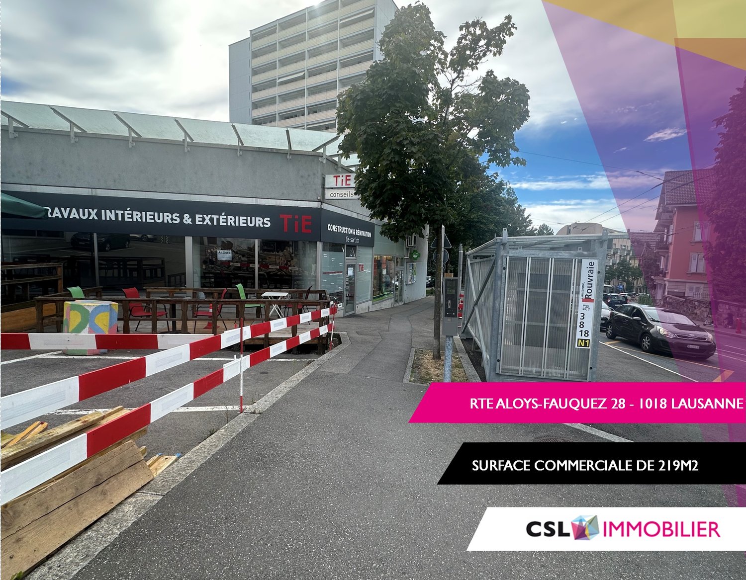 Facade of a commercial building with a sign reading 'ravaux intérieurs & extérieurs Tie', a closed shop with a canopy, a sidewalk, some barriers on the road, parked cars, trees and a building in the background.