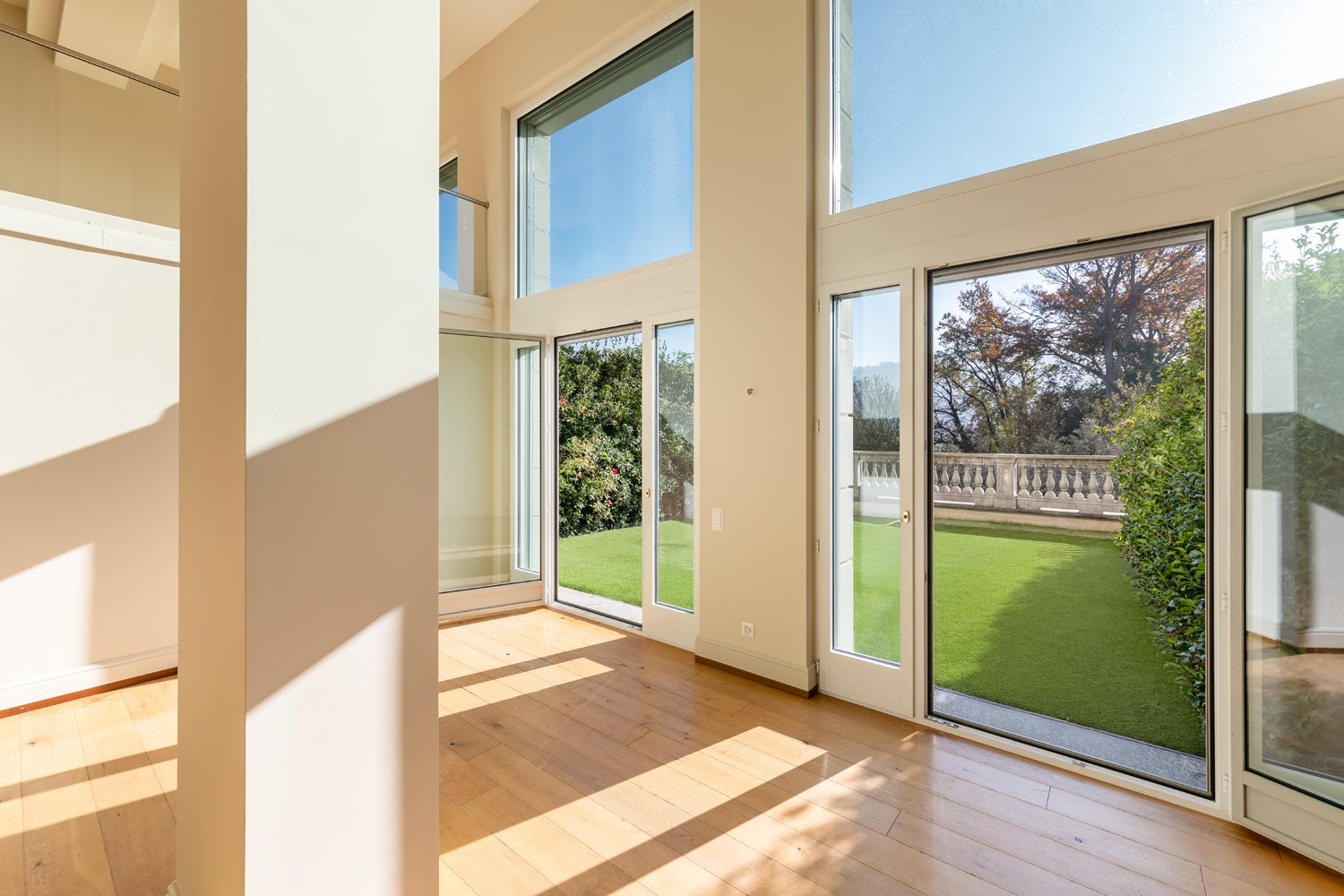 Spacious living room with large windows overlooking a well-manicured lawn and garden. The room features hardwood floors, high ceilings, and an abundance of natural light. The sliding glass doors provide access to the outdoor area.