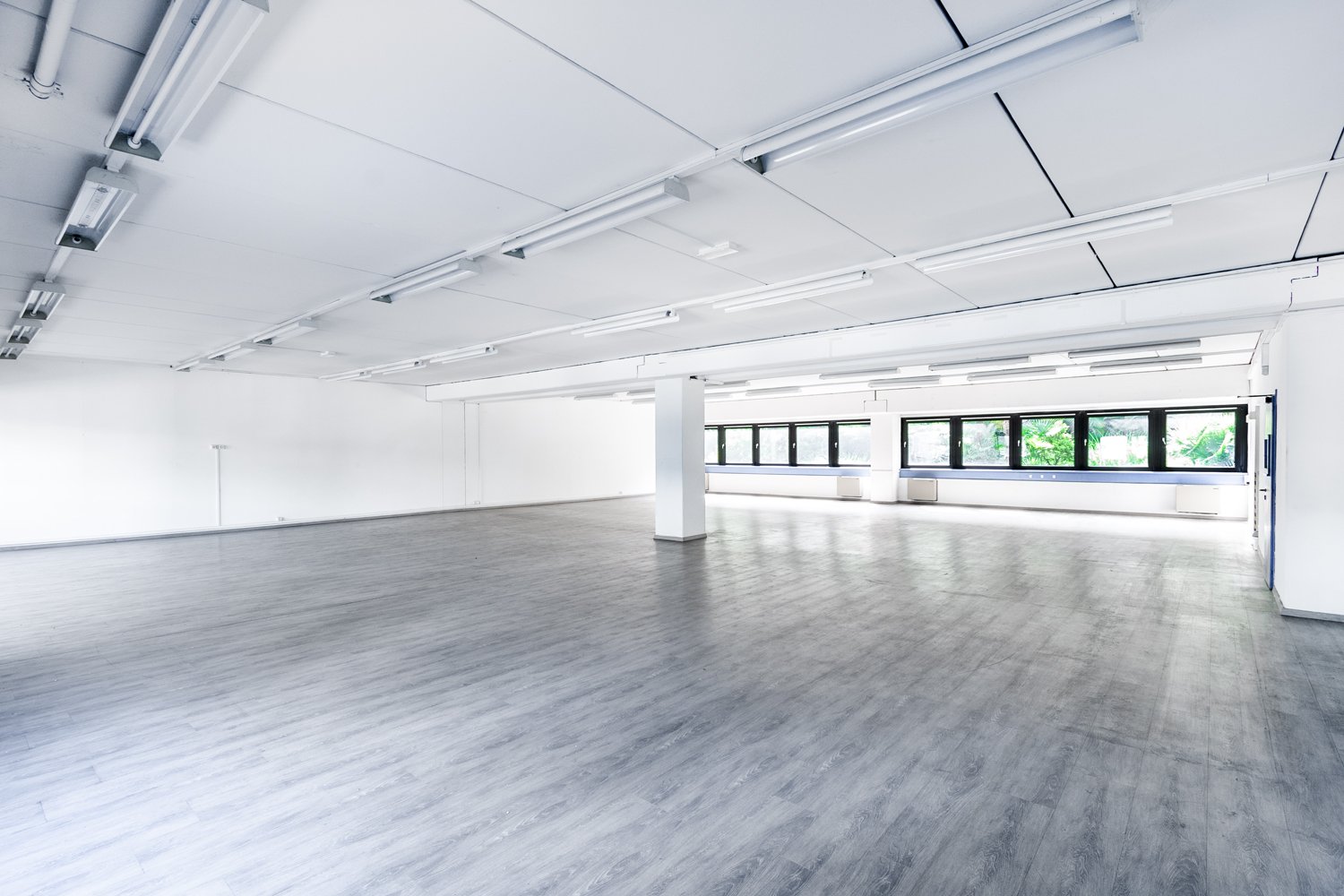 Large open office space with white walls, gray laminate flooring, and overhead fluorescent lighting. The space has large windows along one side providing natural light. The room appears to be empty and ready for use.