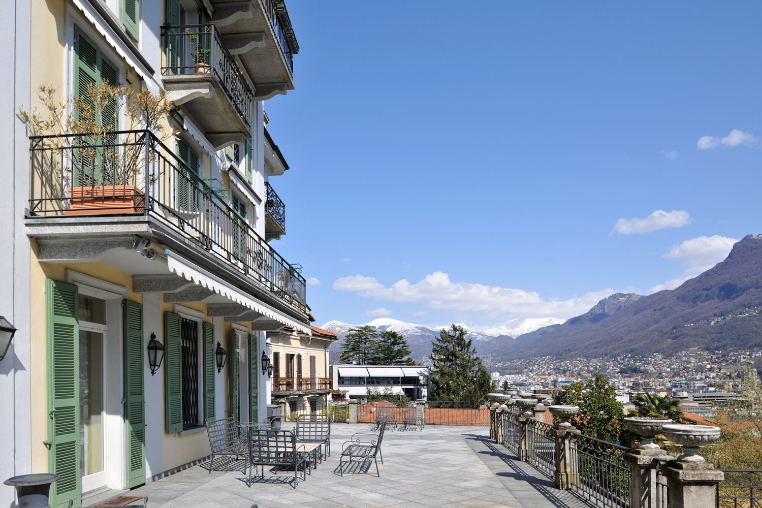 Multi-story house, beige wall, green shutters, metal fence, balcony with plants, patio with chairs and table, mountain and cityscape view
