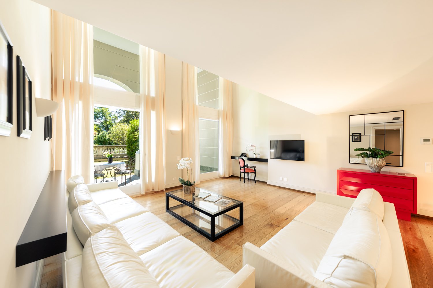 Leather couch, glass coffee table, television, red cabinet, mirror, wooden floor, white walls, picture frames, white curtains, balcony, outdoor furniture