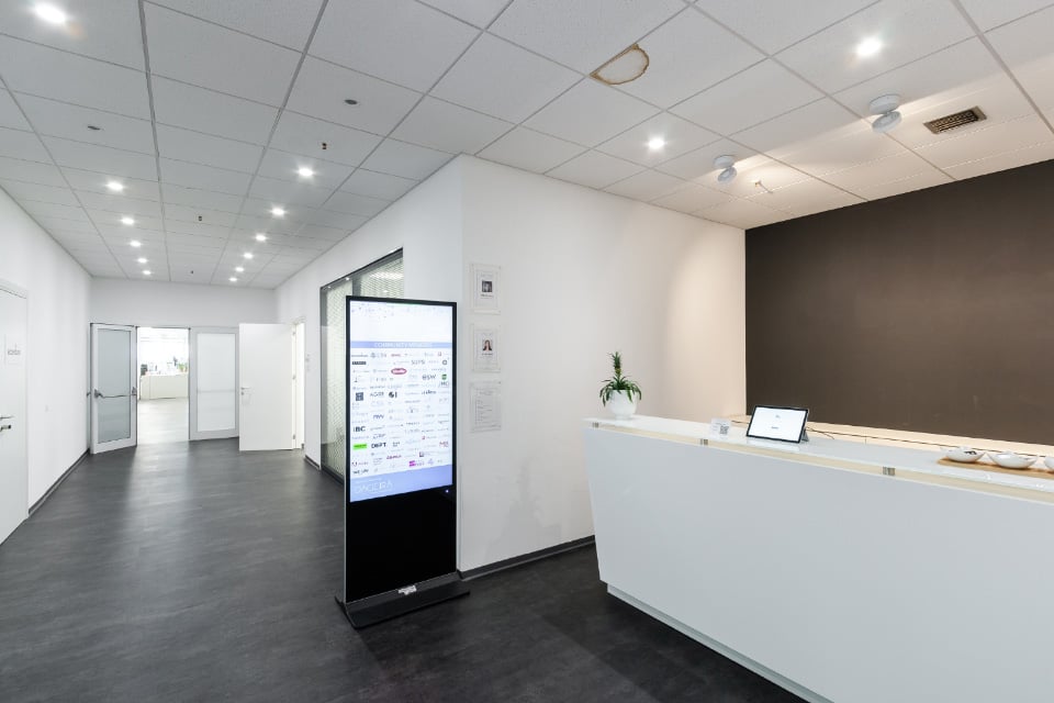 The image shows a modern office space with a reception area. The space has a clean, minimalist design with white walls, a gray floor, and a long white reception desk with a laptop on it. There is a large digital display screen in the center of the room, a