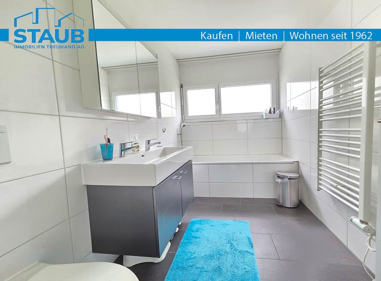 white tiled bathroom, bathtub, sink, toilet, mirror, faucet, soap dispenser, trash can, towel rack, blue rug