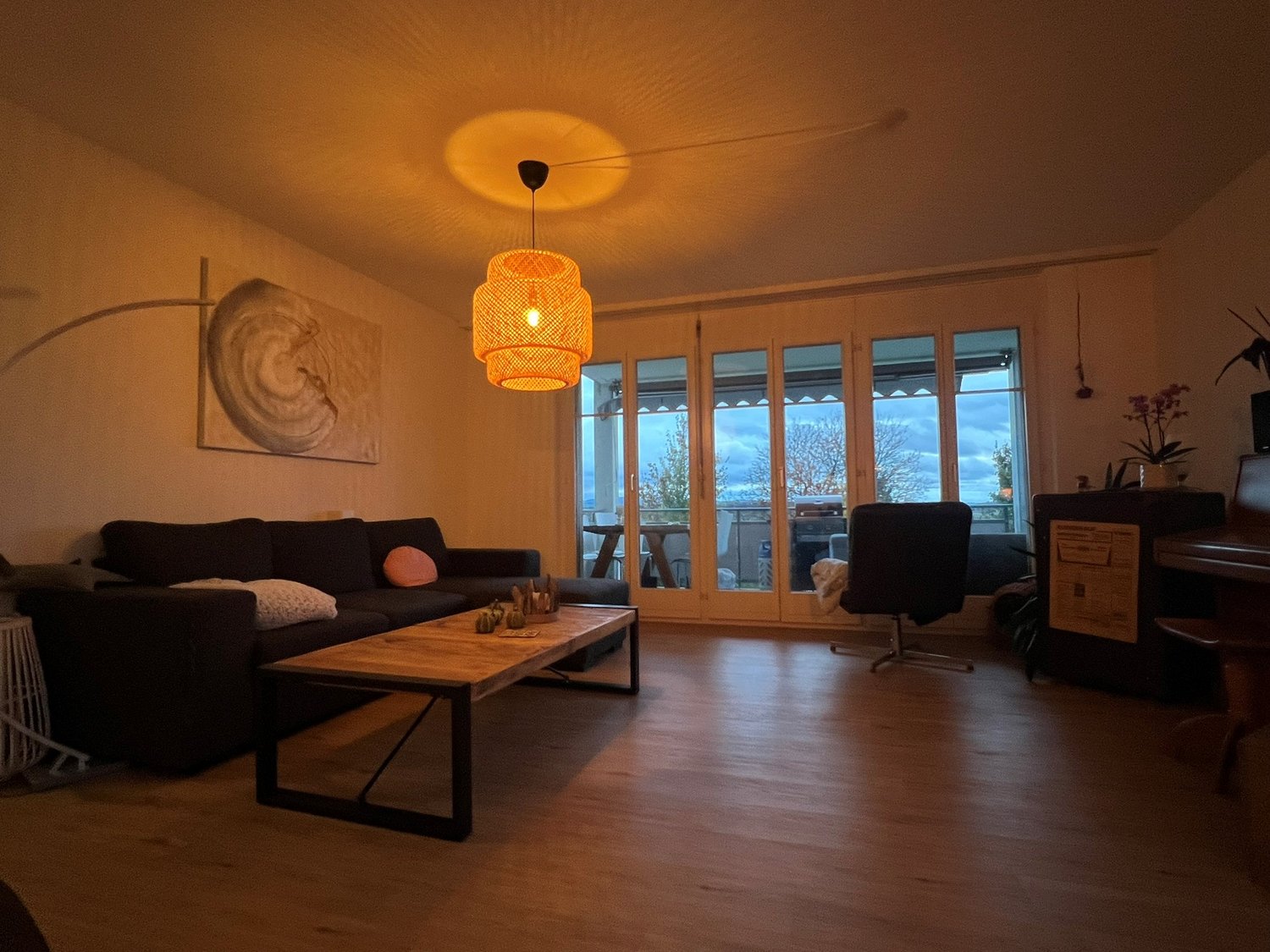 Living room with wooden floor, gray sofa, wooden coffee table, office chair, floor lamp, ceiling lamp, sliding doors with terrace view