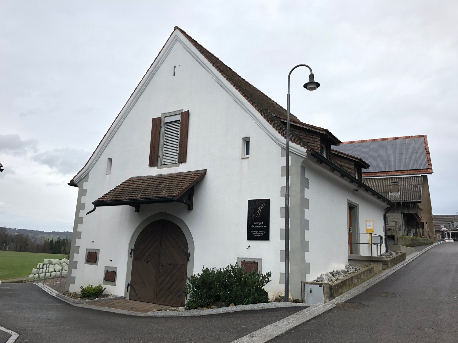 Small white house, brown door, brown roof, solar panels on another building, lamp post, parked car