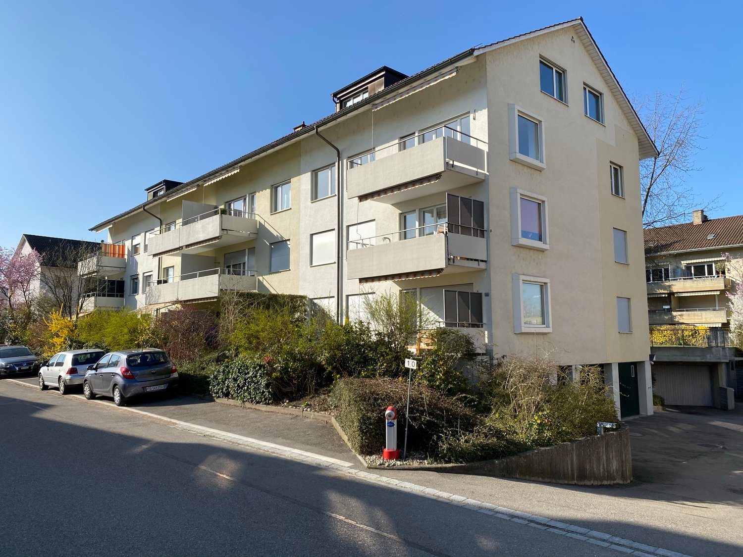 Building with balconies and garage, 4 cars parked outside, shrubs and plants, road with cars