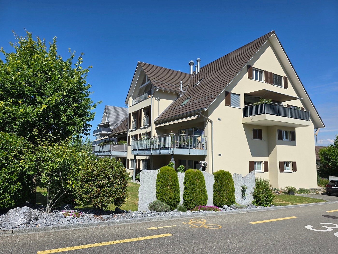 3-story residential building, beige exterior, brown roof, balconies on all floors, small garden, shrubs and trees in front