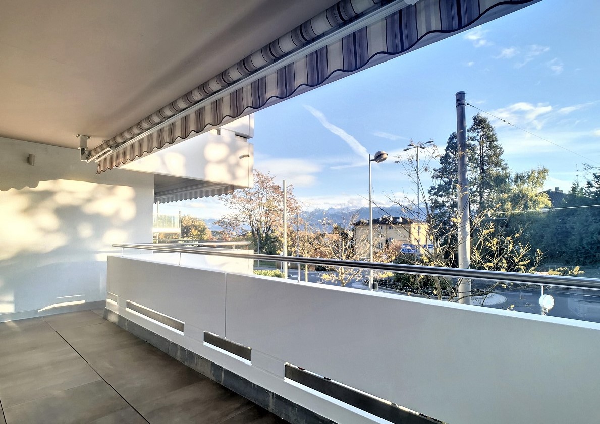 A balcony with a retractable awning, white walls, a metal railing, and a tile floor. The balcony provides a view of the outdoors, with streetlights, trees, and mountains in the distance.