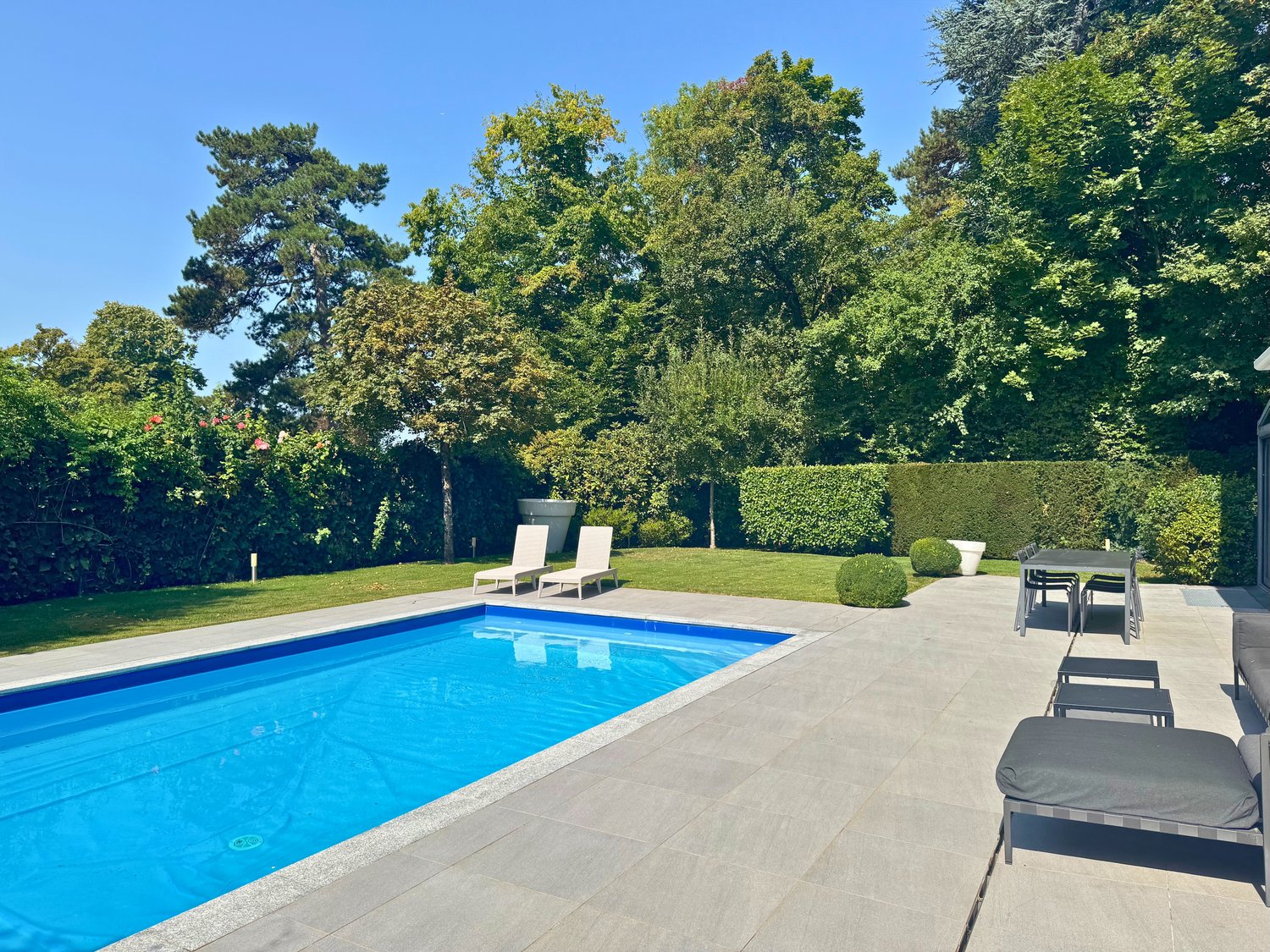 Large garden with a swimming pool, lounge chairs, table and chairs, and landscaped hedges and trees.