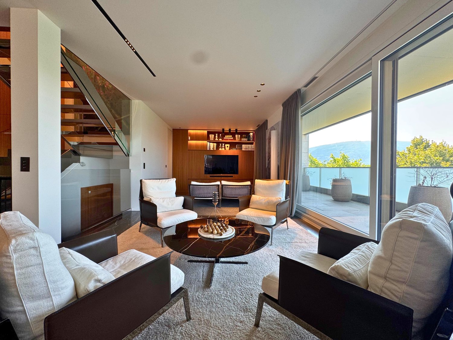 Modern living room with large windows, white chairs, black marble coffee table, and a TV on a wooden shelf.
